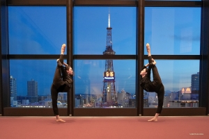 Dua penari menampilkan pose yang mengesankan, meniru puncak Menara Televisi Sapporo yang menjulang tinggi, yang terlihat melalui jendela.
